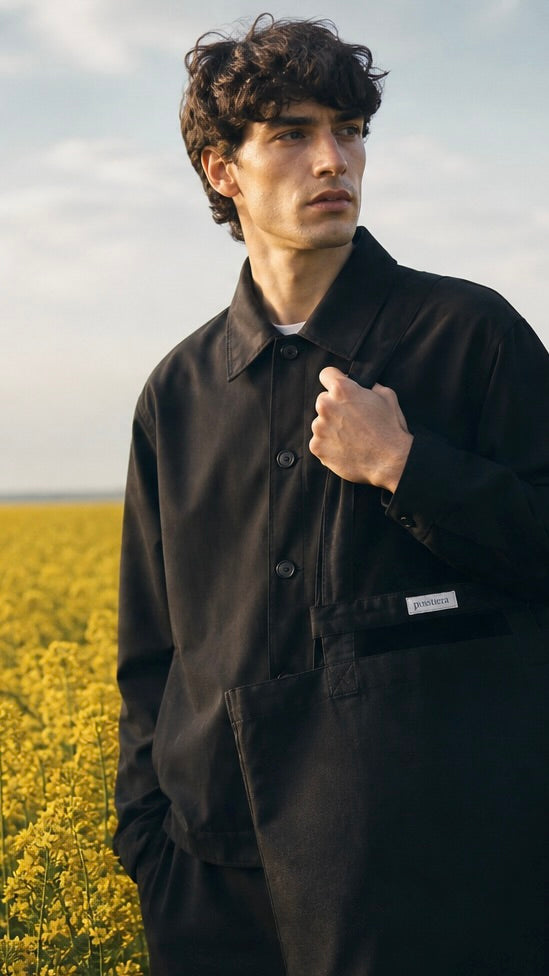 Man wearing a black jacket standing in a field of yellow flowers having a black caviar pusstiera tote bag