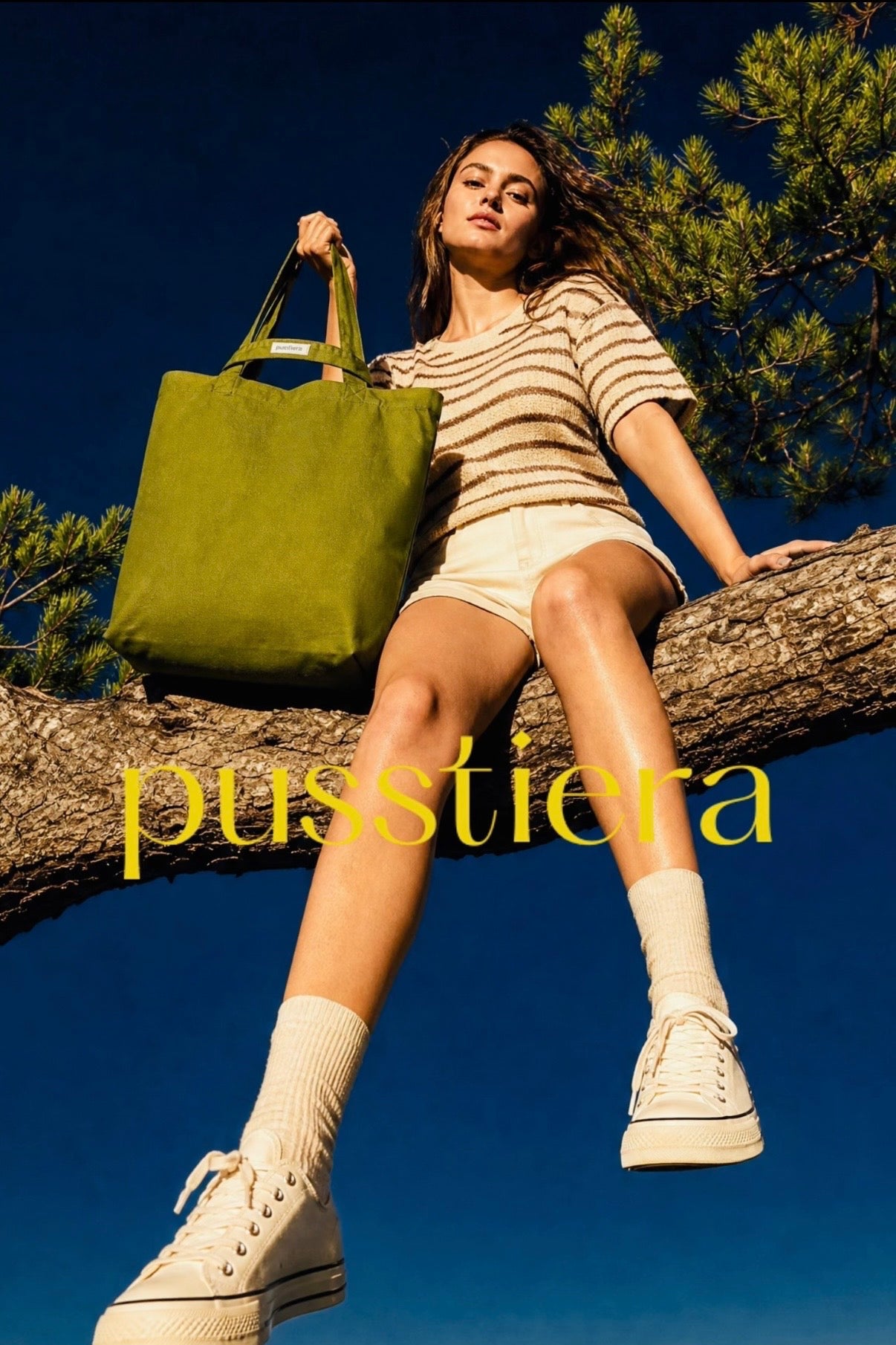 A woman sits on a tree branch outdoors under a blue sky, wearing a beige and white striped short-sleeve top, white shorts, white socks, and white high-top sneakers, while holding a large avocadogreen tote bag. The brand name 'pusstiera' appears in yellow text across the center.